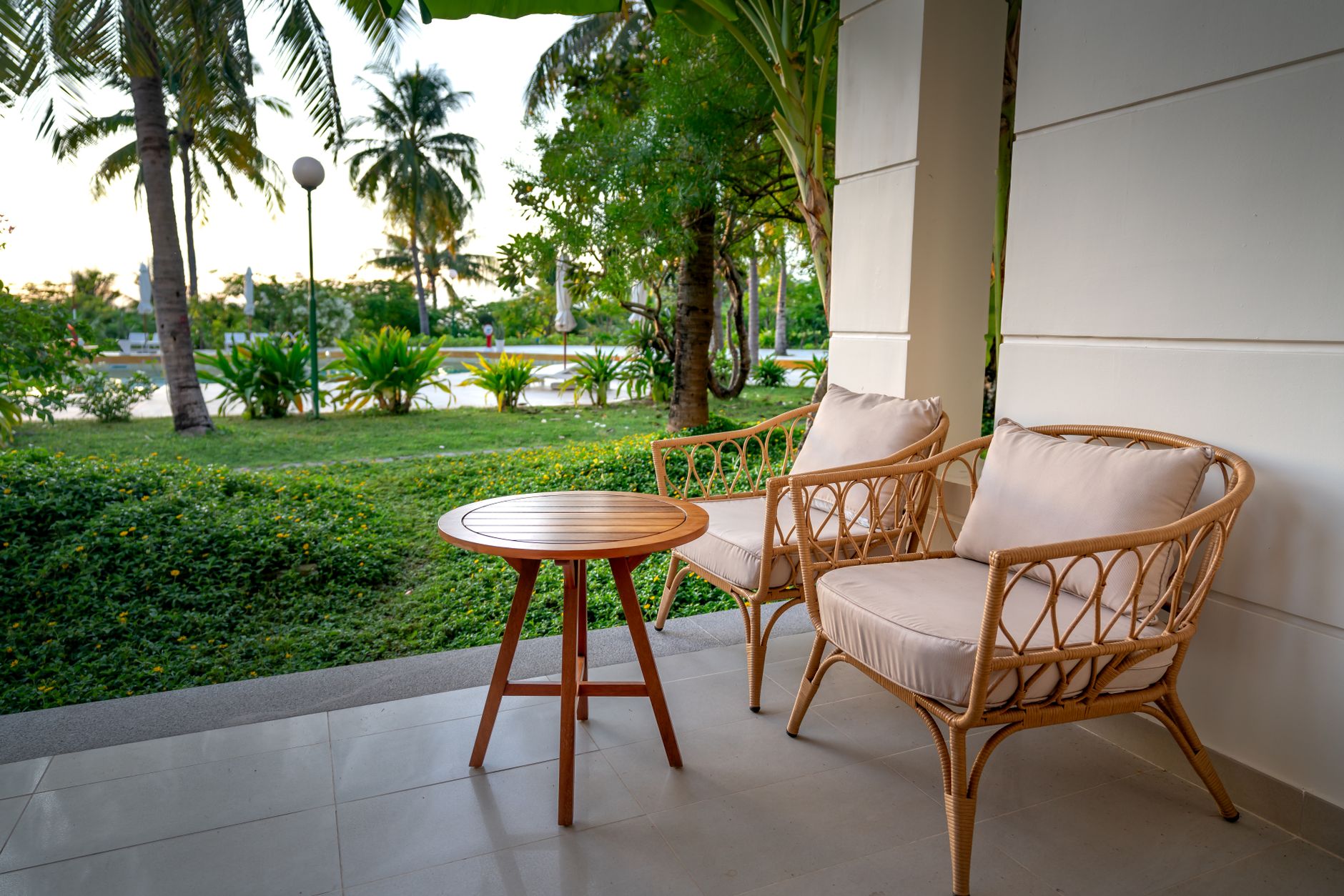 Two rattan chairs and a round timber table on a resort verandah facing a palm garden