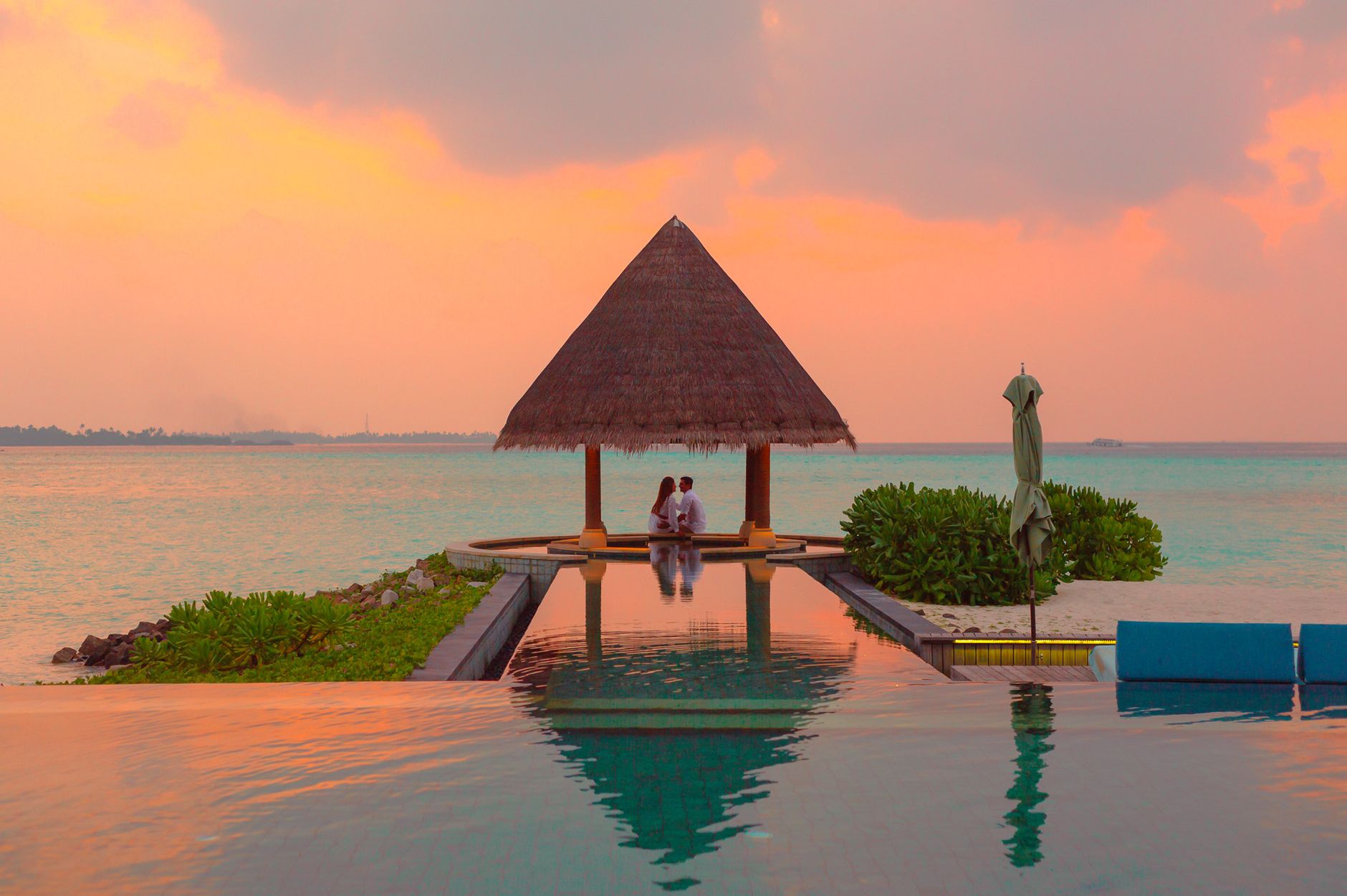 Couple by an infinity pool and thatched pavilion facing the sea at sunset